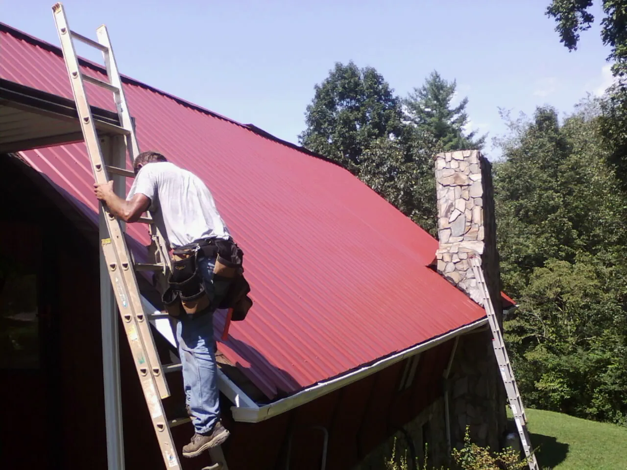 Roofer on a ladder beside a red metal roof and stone chimney — Soffit & Fascia Repair by Roofing Brothers in Hegins